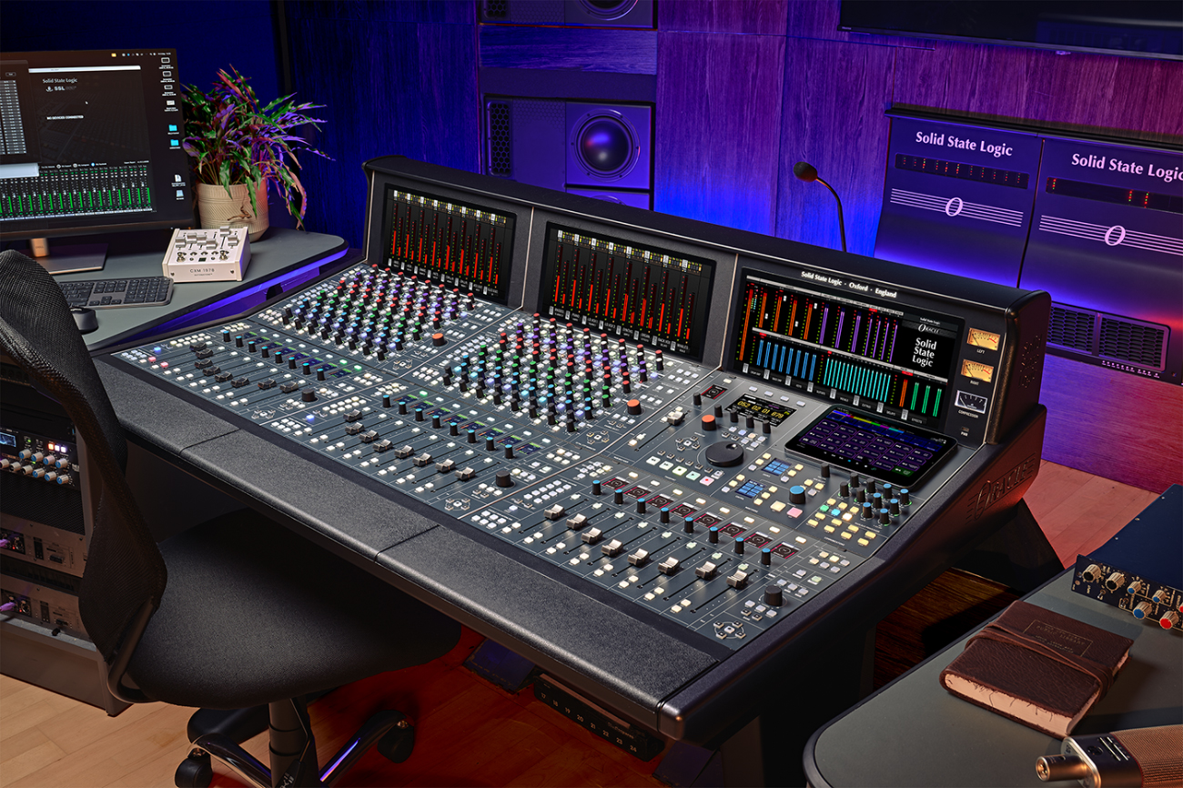 Modern studio with a pro audio mixing console featuring colored knobs, faders, digital screens, and purple-blue accent lighting.