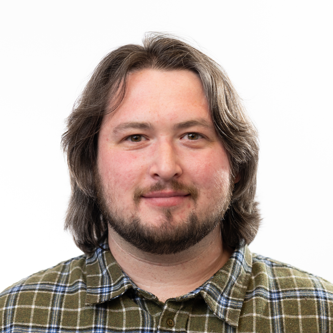 Smiling man with shoulder-length brown hair, beard, and green plaid shirt stands against plain white background.