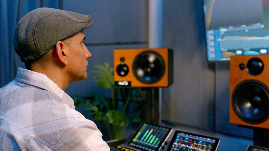 Man in flat cap at recording console with KSD monitors, plaid shirt, speakers, perfecting audio mix in pro studio setting.