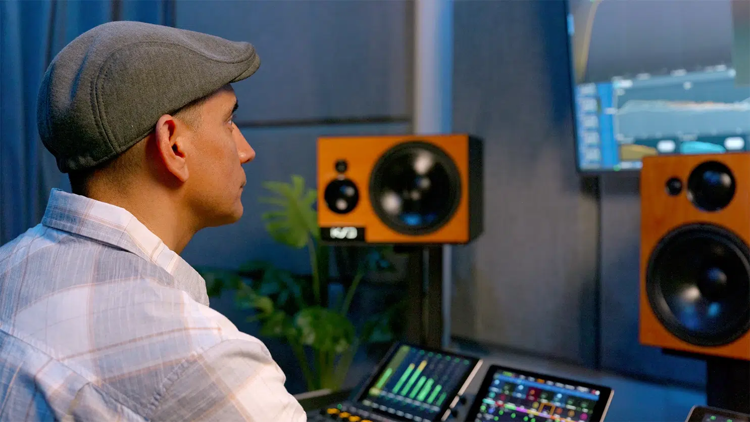 Man in flat cap at recording console with KSD monitors, plaid shirt, speakers, perfecting audio mix in pro studio setting.