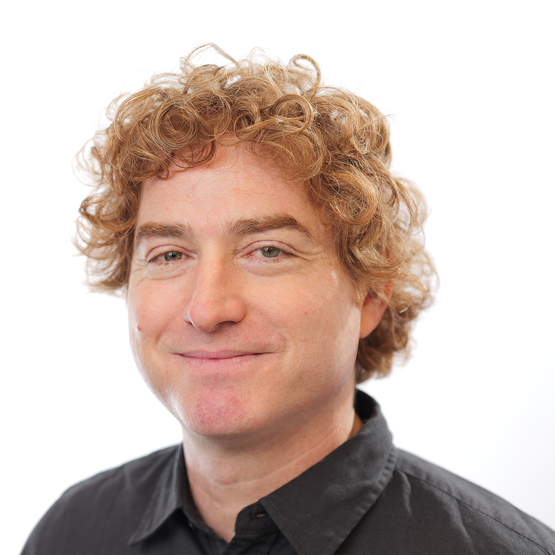 Smiling man with curly light brown hair and fair skin in black collared shirt, facing camera on white background.