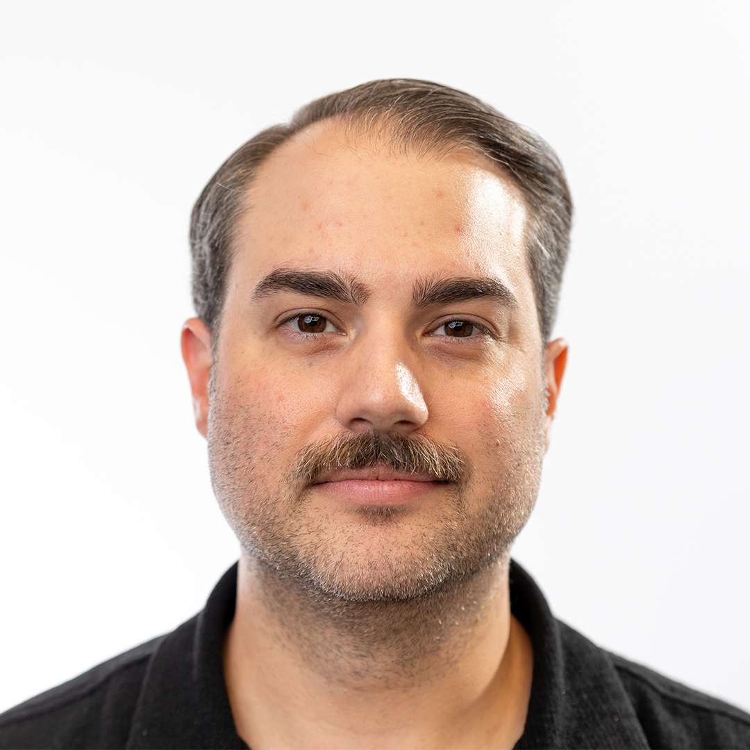 Man with short brown hair, mustache, and light beard in black shirt on white background, facing forward with neutral expression.