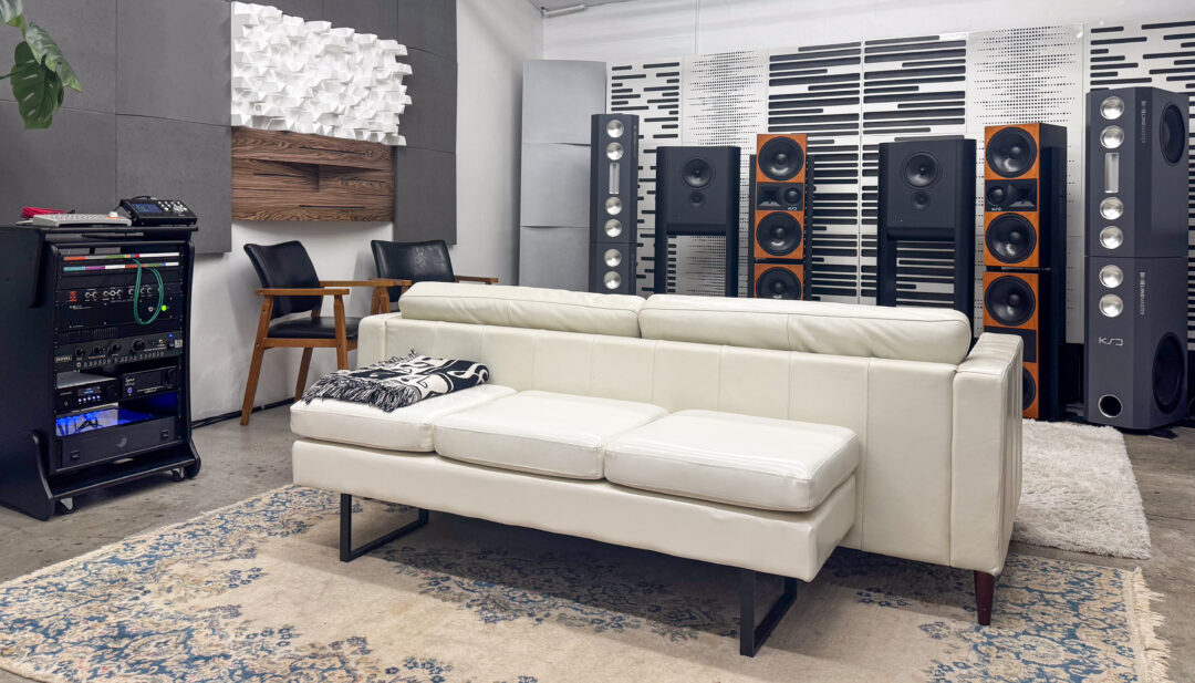 Sleek audio room with white leather sofa, patterned rug, wood chairs, rackmount gear, large black speakers, sound panels.