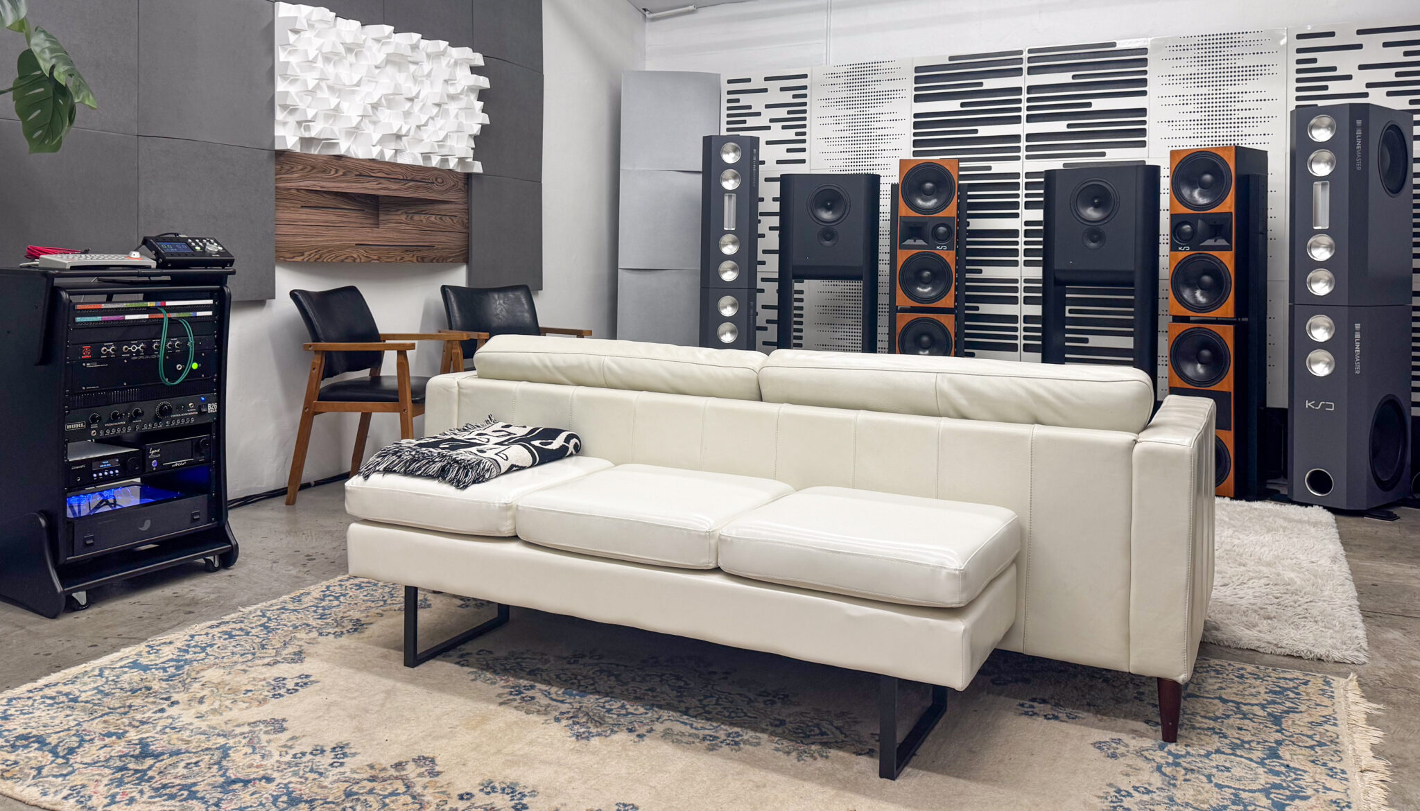 Sleek audio room with white leather sofa, patterned rug, wood chairs, rackmount gear, large black speakers, sound panels.