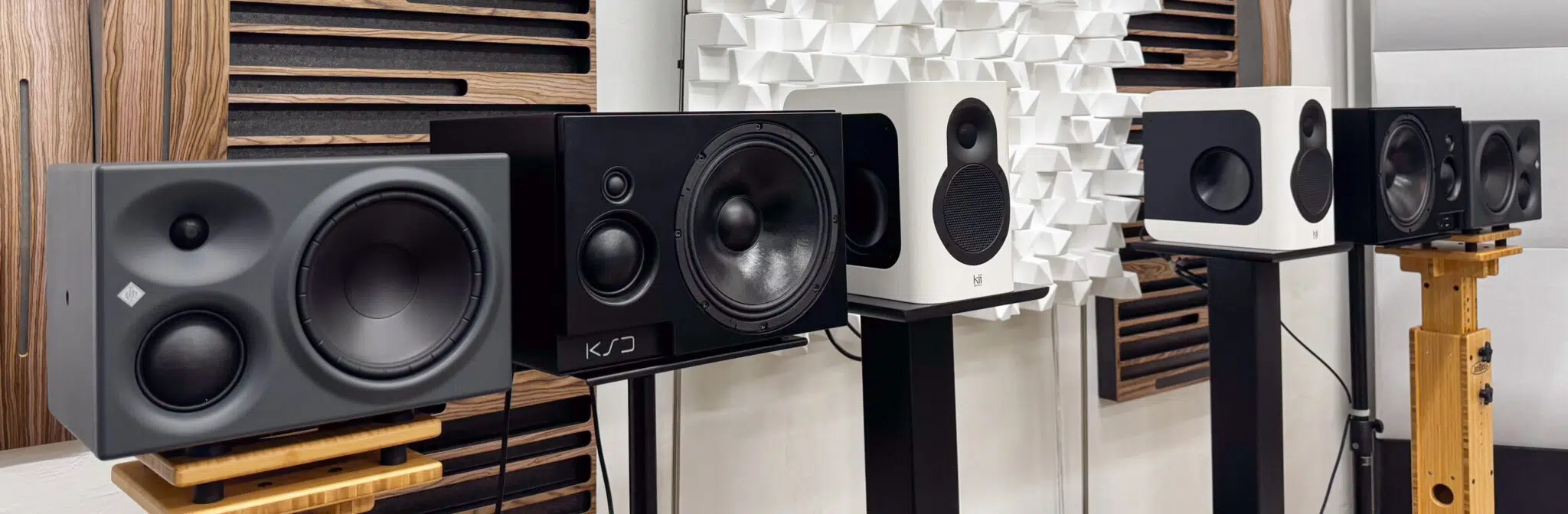 Studio monitor speakers of various sizes and brands on stands in an acoustically treated room with wall panels.