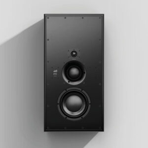 Black 3-way studio monitor with three round drivers, vertically mounted on a light gray wall, soft shadow to the left.